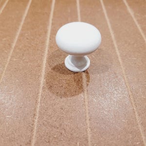 May include: A white, round cabinet knob with a short, pedestal base. The knob is a solid, smooth sphere. The base is a small, circular disc. The knob is set against a light brown, wood-grain surface.