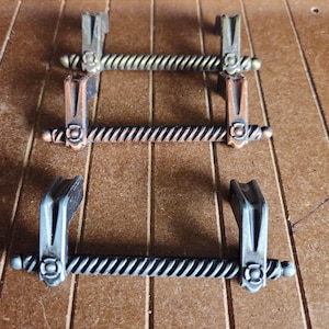 May include: Three decorative metal towel bars in bronze, copper, and silver tones. Each bar has a twisted design and is mounted on decorative brackets. The bars are arranged on a wooden surface.