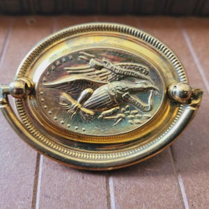 May include: Gold-toned oval cabinet knob with a raised design of an eagle holding a bundle of arrows and an olive branch. The eagle is holding a banner that reads "E Pluribus Unum".