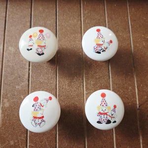 May include: Four white ceramic cabinet knobs with a colorful clown design. Each knob features a different clown illustration with red, blue, and yellow accents. The knobs are round and have a glossy finish.