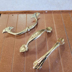 May include: Three gold-colored cabinet pulls in various designs. The pulls are made of metal and have a shiny finish. One pull is curved, another is a bar shape, and the third has a decorative end. They are displayed on a brown surface.