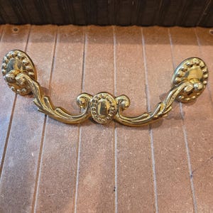 May include: A gold-colored metal drawer pull featuring a swinging handle with ornate leaf-like detailing and a central oval medallion. The handle is attached to two circular mounting plates, each decorated with a beaded border along the outer edge.