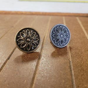 May include: Two decorative metal knobs with a floral design. One knob is bronze-colored, and the other is a dark gray. The knobs are round with a raised flower pattern in the center. The background is a wooden surface.