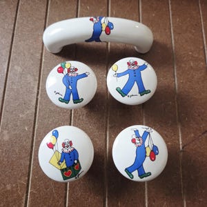 May include: A set of white ceramic cabinet knobs and a handle featuring a whimsical clown design. The clowns are depicted in blue suits with red and white accents, holding balloons. The knobs and handle are set against a brown wooden surface.