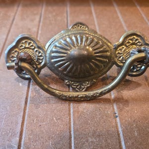 May include: A brass-toned metal drawer pull featuring a central oval plate with a sunburst pattern and two side plates with intricate floral cutouts. A curved, textured handle is attached to the side plates, showing signs of oxidation.