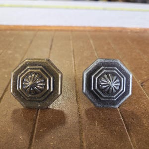 May include: Two octagonal cabinet knobs. One is bronze-colored, the other is silver-toned. Each knob features a central starburst design within a series of concentric octagons. The knobs are likely for furniture or decorative use.