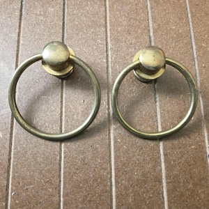May include: Two vintage brass ring pulls with a circular design and a small, round backplate. The pulls are set against a light brown wooden surface. These hardware pieces are likely for cabinets or drawers.