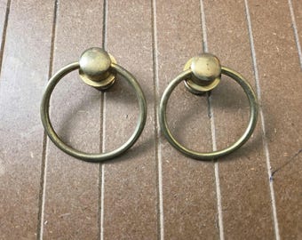 May include: Two vintage brass ring pulls with a circular design and a small, round backplate. The pulls are set against a light brown wooden surface. These hardware pieces are likely for cabinets or drawers.