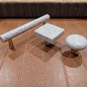 May include: Three cabinet knobs made of white speckled stone with gold metal bases. The knobs are in the shape of a rectangle, a square, and a circle.