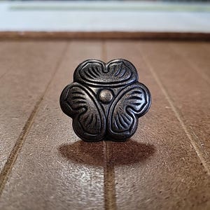 May include: An antique-style cabinet knob with a floral design. The knob is made of a dark, metallic material and features a three-petal design with detailed leaf-like patterns. The knob is approximately 3.8 cm in diameter.