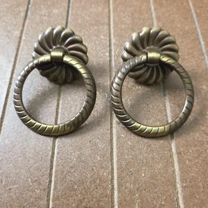 May include: Two antique brass drawer pulls. Each pull features a decorative, ridged ring handle attached to a floral-shaped backplate. The hardware has a warm, aged finish, suggesting vintage or retro design elements. Ideal for furniture restoration or adding a classic touch.