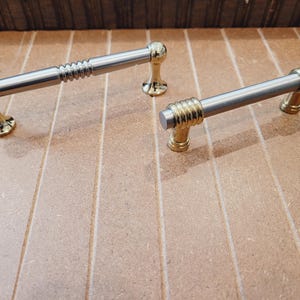 May include: Two gold and silver cabinet door pulls with a decorative design. The pulls are on a wood surface.