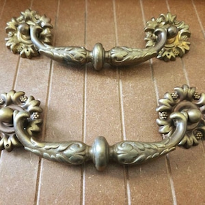 May include: Two antique brass drawer pulls. Each pull features a floral design with a central handle and decorative end pieces. The pulls are a warm gold color and appear to be made of metal.