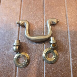 May include: A brass-colored metal furniture handle featuring a curved bail pull and two circular mounting plates with cylindrical posts. The hardware has a smooth, metallic finish and consists of three separate pieces designed for attachment to a drawer or cabinet front.