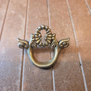 May include: A brass-colored metal drawer pull featuring a central keyhole escutcheon surrounded by a decorative scalloped border and two symmetrical side arms that extend outward, ending in rounded circular accents above a curved handle loop.