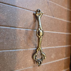 May include: A vertical brass-toned metal drawer pull featuring an elongated, ribbed central body with a smooth oval accent. The handle terminates in ornate, symmetrical decorative flourishes at both the top and bottom ends, with a keyhole cutout.