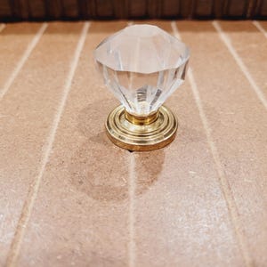 May include: A clear glass diamond-shaped knob with a gold metal base. The knob is a decorative hardware piece.