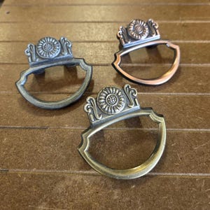May include: Three decorative drawer pulls with a floral design. The pulls have a shield-like shape with a decorative top. One is silver, one is copper, and one is brass. The pulls are on a brown wooden surface.