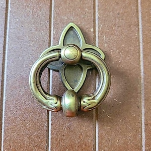 May include: A vintage-style brass door knocker. The knocker features a circular handle attached to a decorative backplate with a fleur-de-lis design. The brass has an aged appearance, suggesting it is an antique or a reproduction.