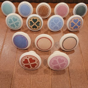 May include: Assortment of 14 ceramic drawer knobs with various designs. Some knobs feature a four-leaf clover design with hearts, while others have a polka dot pattern. The knobs are in various colors, including blue, pink, green, brown, and red.