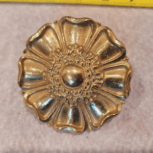 May include: A gold-colored, flower-shaped brooch. The brooch has a detailed design with petal-like edges and a central, raised circular element. The metal has a shiny finish, reflecting light. The brooch is likely vintage or antique.