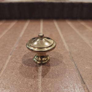 May include: A small, gold-colored decorative knob with a rounded top and a pedestal base. The knob has a shiny finish and is likely made of brass or a similar metal. It is designed for use on furniture or cabinetry.