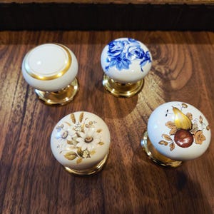May include: Four decorative ceramic cabinet knobs. Each knob features a different design: a white knob with a gold band, a blue floral pattern, a floral design with daisies, and a fruit design. All knobs have a gold-colored base.