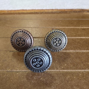 May include: Three decorative cabinet knobs with a circular design. The knobs have a raised center with smaller circles and are surrounded by concentric rings. The knobs are in shades of bronze, silver, and gold.