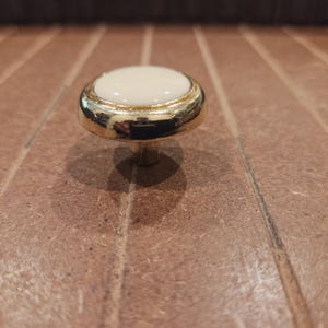 May include: A cabinet knob with a gold-coloured base and a round, off-white centre. The knob has a shiny finish and is set against a brown, textured surface. This hardware is suitable for drawers and cabinets.