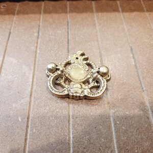 May include: Ornate brass drawer pull with a detailed, symmetrical design. The pull features a central medallion shape, flanked by curved handles and decorative embellishments. The brass has a warm, aged patina.