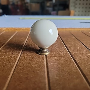 May include: A white ceramic knob with a brass base. The knob is round and has a smooth finish.