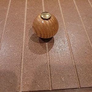 May include: A round wooden knob with a gold metal top. The knob is made of light brown wood with a grain pattern.