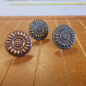 May include: Three decorative cabinet knobs with a sunflower design. The knobs are made of metal and have a bronze, copper, and antique brass finish. The knobs are round and have a raised floral pattern.