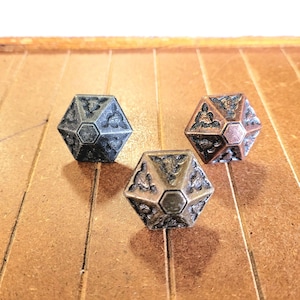 May include: Three decorative metal dice with intricate geometric designs. The dice are in shades of gray, bronze, and copper, with a detailed pattern on each face. They are arranged on a wooden surface.
