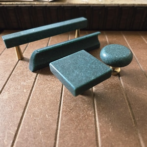 May include: A collection of dark green cabinet hardware, including two rectangular pulls, a curved pull, a square knob, and a round knob. The hardware has a speckled texture and is mounted on brass bases.