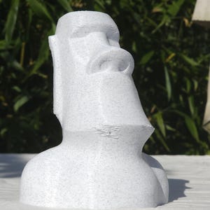 May include: A white Moai statue, a sculpture of a large human face and torso, with a prominent nose and chin. The statue is made of a speckled material and is set against a blurred green background.