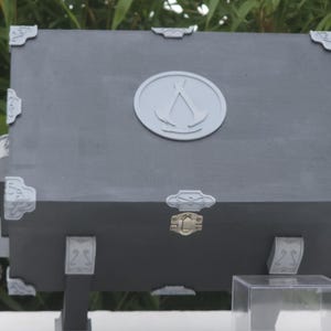 May include: A grey wooden chest with decorative silver corner accents and a circular emblem on the front. The chest has a gold-coloured clasp and is supported by black legs. A clear plastic box is in front.