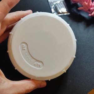 Peut inclure: Un couvercle blanc et circulaire avec les mots "AUF" et "ZU" en relief sur la surface. Le couvercle comporte une série de petites arêtes en relief sur le bord. Le couvercle est tenu par une main.