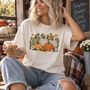 Puede incluir: Una camiseta de color crema con un estampado gráfico de cuatro calabazas en tonos verde, blanco y naranja, rodeadas de flores y corazones pequeños. La camiseta se combina con unos vaqueros de mezclilla azul claro.