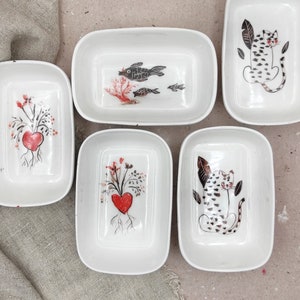 Könnte beinhalten: Eine Sammlung weißer rechteckiger Schalen mit handgemalten Designs. Die Designs umfassen ein rotes Herz mit Wurzeln und floralen Elementen sowie Illustrationen von Fischen und Leoparden in Schwarz und Weiß.
