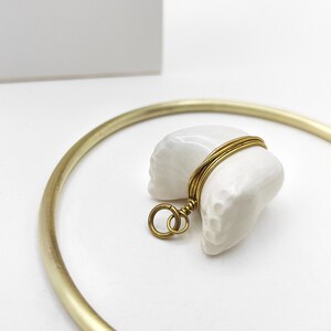 Könnte beinhalten: Weißer Porzellan-Glückskeks-Anhänger mit einem goldenen Draht umwickelt. Der Anhänger hat eine Schlaufe zum Anbringen an einer Halskette oder einem Armband.