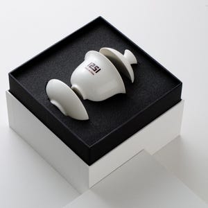 May include: A white ceramic tea set, including a cup, lid, and two other pieces, presented in a black box with a white outer box. The cup has the text "1251 Made in Chine" printed on it. The set is displayed against a neutral background.