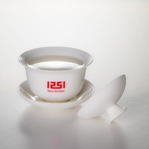 May include: A white porcelain teacup and saucer set with the text "1251 Blanc de Chine" in red. The teacup is filled with a clear liquid. A matching lid rests beside the cup and saucer.