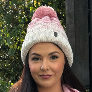 May include: A cream and pink cable-knit beanie hat with a matching pom-pom. The hat has a small logo on the side. A warm winter accessory for cold weather.