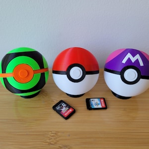Puede incluir: Tres objetos coloridos con temática de Poké Ball. Uno es verde y negro, otro es rojo y blanco, y el tercero es morado y blanco con una "W" blanca. Dos pequeños cartuchos de juego están frente a las bolas.