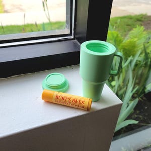 May include: A light green insulated tumbler with a handle, a matching lid, and a tube of Burt's Bees Beeswax Lip Balm. The lip balm tube is orange with yellow and red text. The tumbler and lid are on a white surface.