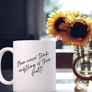 Peut inclure: Mug en céramique blanche avec l'inscription "Men never think anything is their fault!" en écriture noire. Le mug est à côté d'un vase de tournesols. La anse de la tasse est sur le côté gauche.