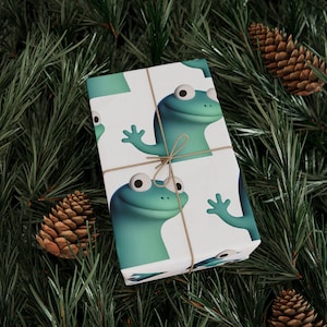 Puede incluir: Papel de regalo con un patrón repetido de ranas de dibujos animados en tonos verdes y azules sobre un fondo blanco. El paquete envuelto está atado con hilo y descansa sobre un lecho de agujas de pino y piñas.