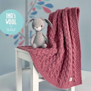 May include: A pink knitted baby blanket with a cable knit pattern, draped over a white chair. A gray stuffed bunny sits on the blanket. The blanket has a label that says "INA'S WOOL THE WAY OF KNITTING".