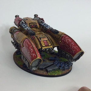 May include: A gold and red miniature tank with a detailed paint job. The tank has two large cannons on the top and is sitting on a base with green grass and rocks.